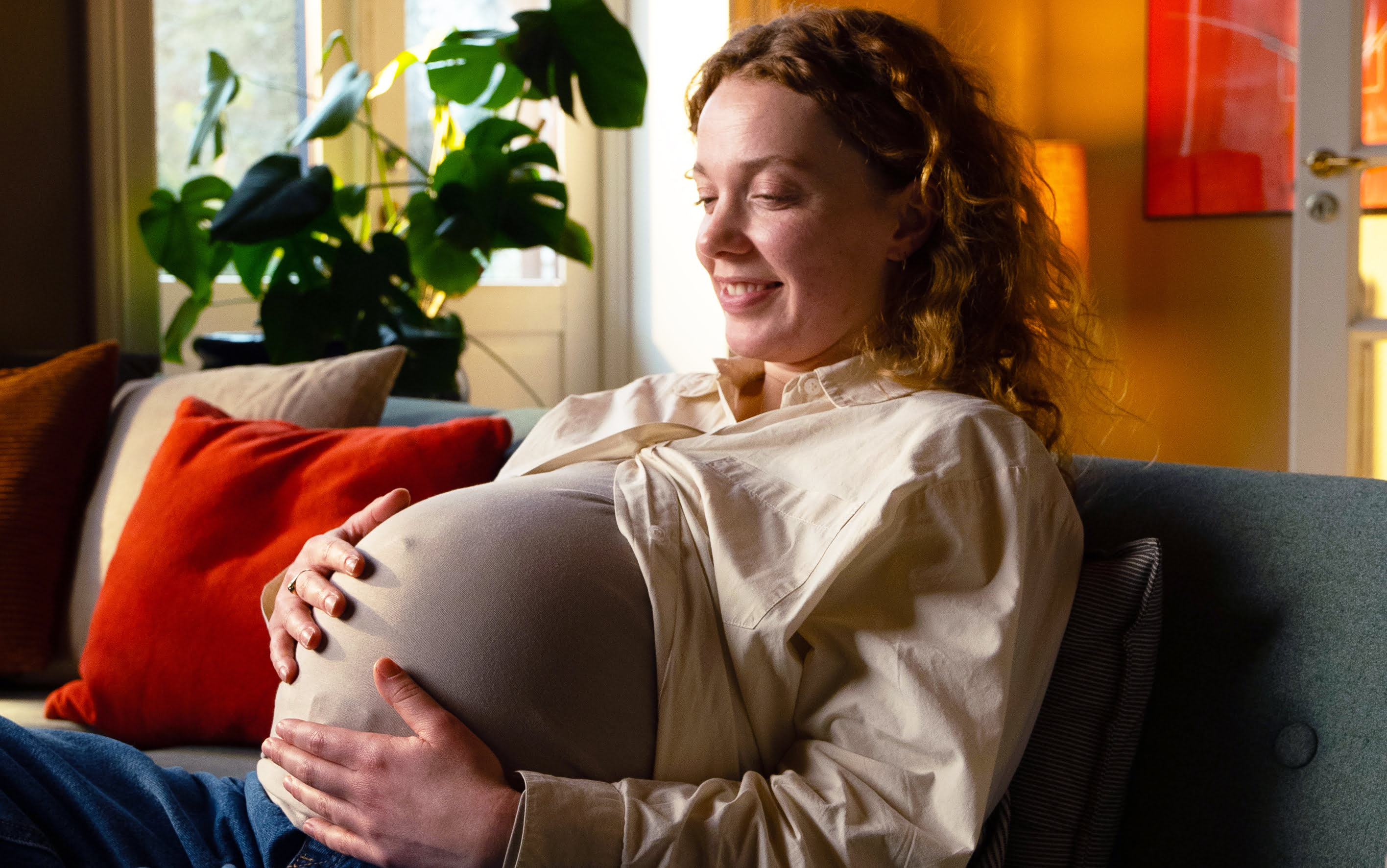Gravid kvinde sidder i sin sofa og smiler imens hun holder sin mave