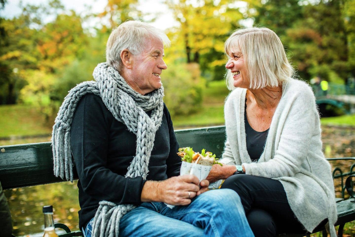 ældre par sidder i på en bænk i en park og smiler til hinanden